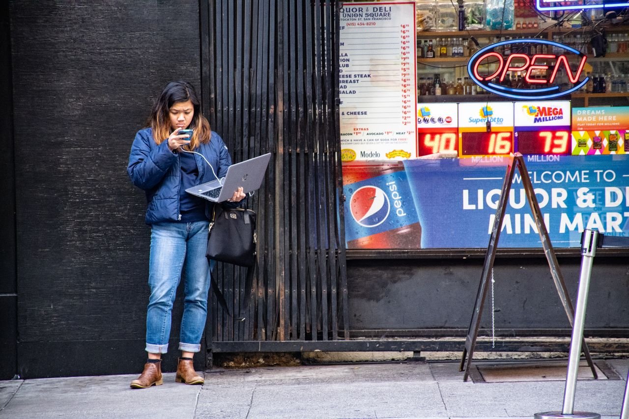 services-03 Asian woman using a laptop and phone outside a city store by an open sign.