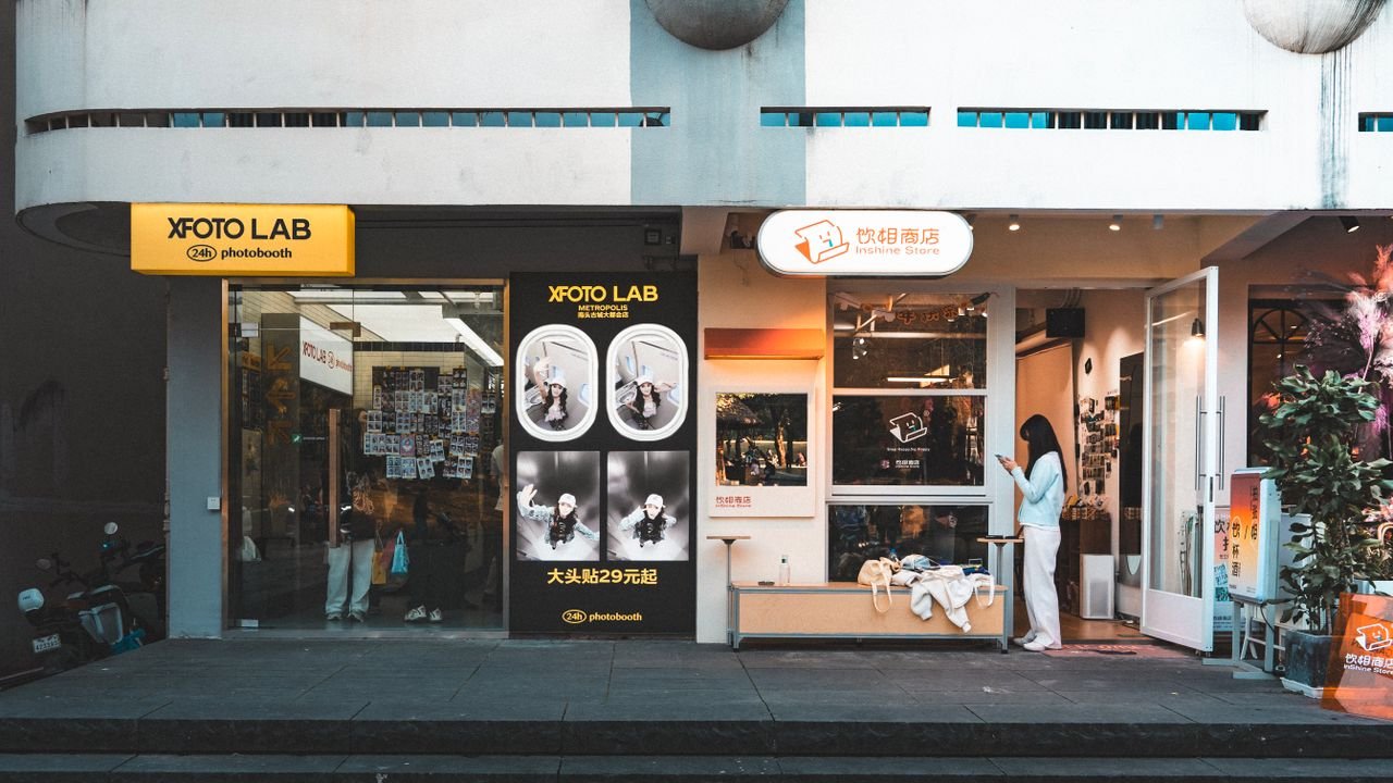 who-we-are Street view of a modern photo lab storefront with a customer using a smartphone.